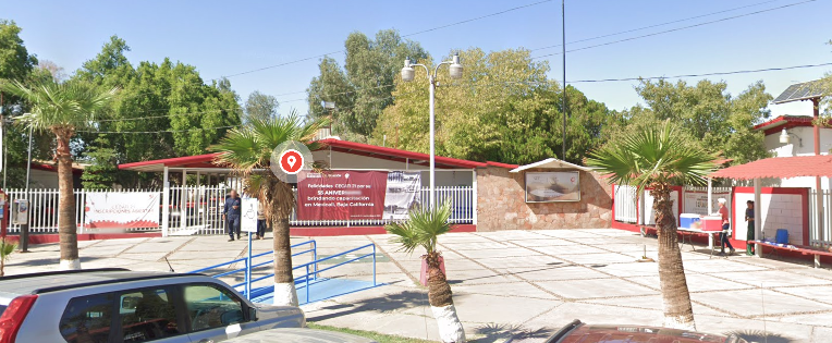 En la fotografía se muestra una pequeña explanada a la que se puede ingresar por medio de una rampa. Al fondo se puede observar la entrada conformada por una reja color blanco y una lona con los datos del plantel.